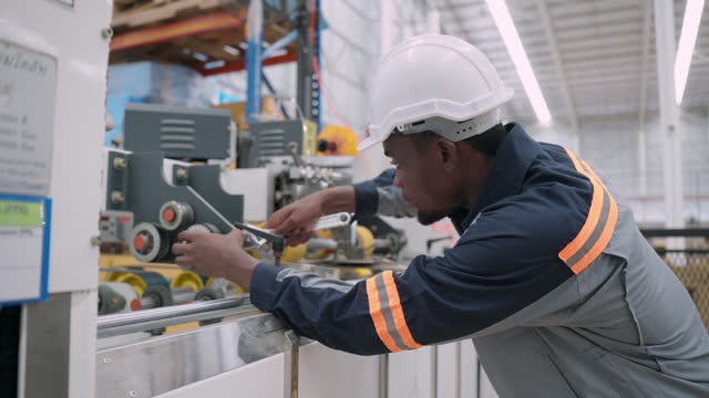 African male maintenance engineer with protective workwear using wrench to repair the manufacturing machine in the paper production factory. Young adult male technician working with the paper finishing production line.