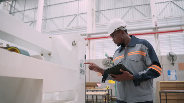 Young adult African engineer wearing protective workwear examining the specification of the paper product manufacturing machine to match with the document in a digital tablet before starting the production line.