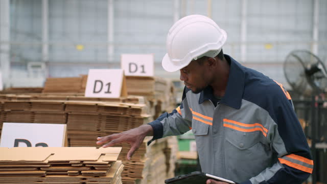 Black adult quality control officer with protective workwear examining the cardboard products quality on a pallet at a warehouse and checking the specification using digital tablet.