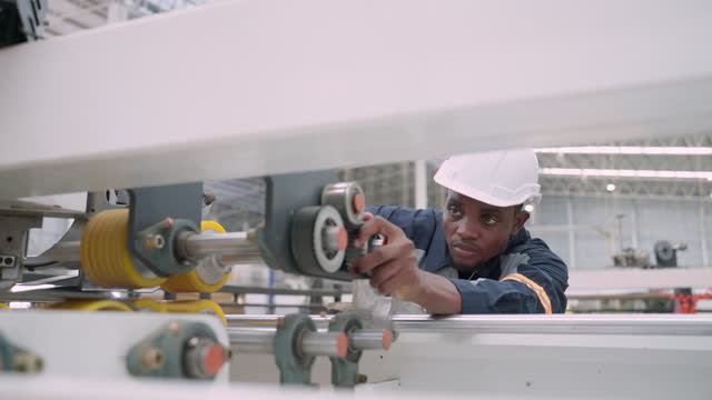 Close up shot of black male engineer with protective workwear adjusting the position of the roll of the manufacturing machine. African male inspecting the machinery of the production line of paper factory.