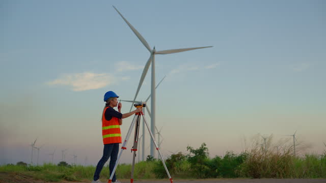 A young technician looks through a theodolite to level land for a wind turbine base in a power generation area.