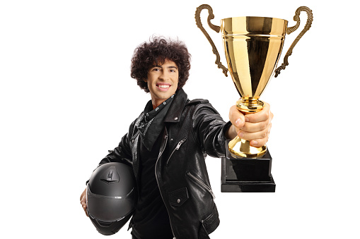 Guy in a black leather jacket holding a helmet and a gold tropy cup isolated on white background