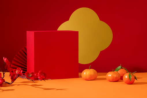 On the table, there are tangerines, peach blossom branches, a gift box and a paper fan. Orange-red background with a holiday theme. Empty space for product display with front view.