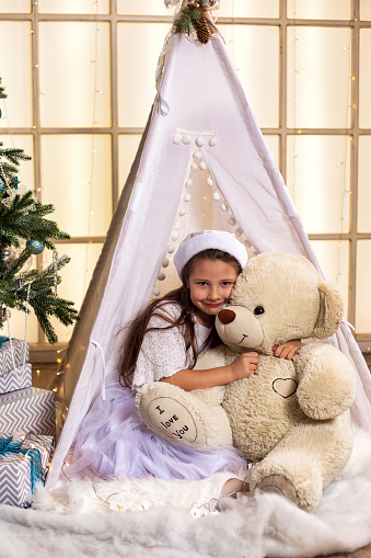 Child girl hugging teddy bear sitting in wigwam near Christmas tree, happy looking at camera. Smile kid girl 5 year old in white dress in children room. Christmas magic concept. Copy ad text space