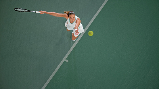 Overhead view of female tennis player serving tennis ball in tennis court.