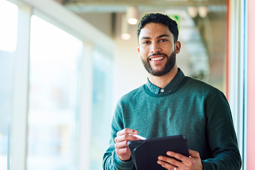Bearded young businessman holding digital tablet, smiling cheerfully at camera in office
