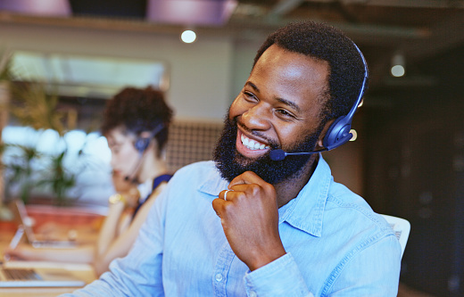 Bearded consultant wearing headset listening attentively to customer with cheerful smile and looking into distance in call center with colleague in background