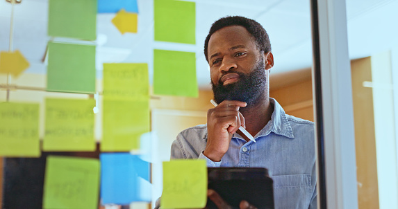 Businessman holding stylus and digital tablet, touching beard and planning work strategies with sticky notes on glass wall in office