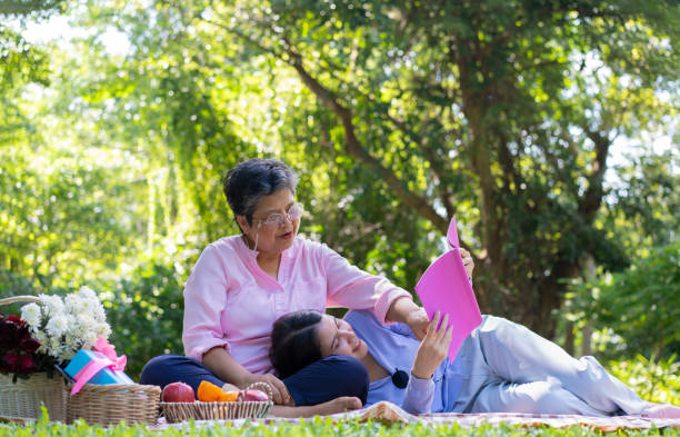 asian daughter sleeping on mother lap and reading book on picnic mat in the park. a happy senior woman talks with her daughter. concept of healthcare and elderly caregiver support service. - grandparent senior adult child reading ストックフォトと画像
