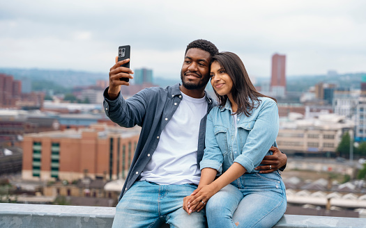 A handsome man and a beautiful woman sitting on the barrier, taking selfies, hugging, talking in front of a modern urban city background dreaming about the future