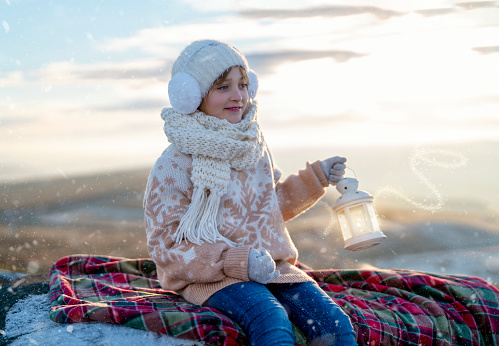 Teenager girl in Christmas sweater sitting on top of a mountain  with lantern with magical light on cold winter day