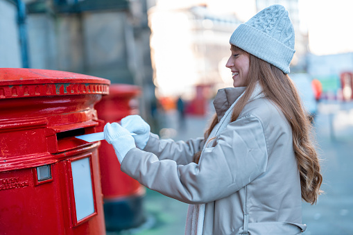 woman in a coat and white hat  putting a card in the red postbox and walking around an English city on a snowy day