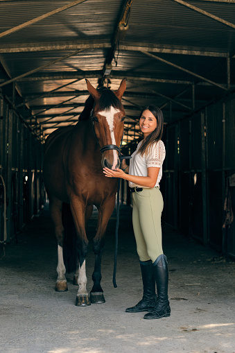 Delighted horsewoman standing with chestnut obedient horse in barn in countryside and looking at camera