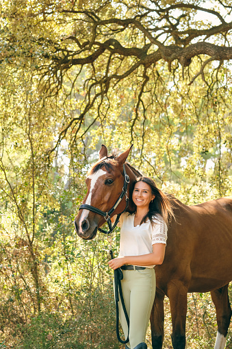 Delighted female equestrian hugging brown horse while standing in green summer woods and looking at camera