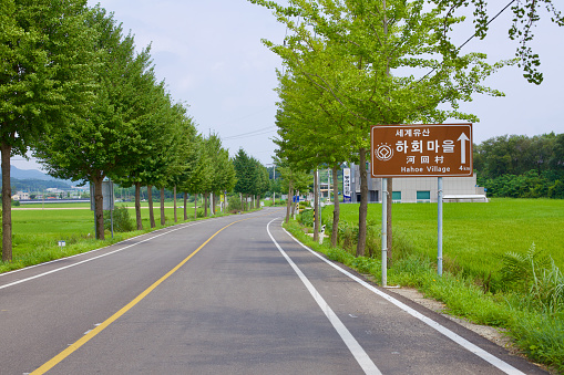 Andong City, South Korea - August 12th, 2023: A serene two-lane, tree-lined road near Hahoe Village, featuring a sign pointing towards the village and a modest shoulder accommodating bike riders, encapsulates the rural charm of the area.