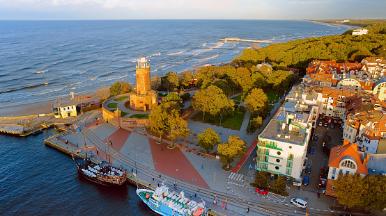 Drone photo captures Kolobrzeg's maritime charm, featuring the iconic lighthouse, cerulean sea, turbulent waves, a distant pier, and autumnal hues on the trees.