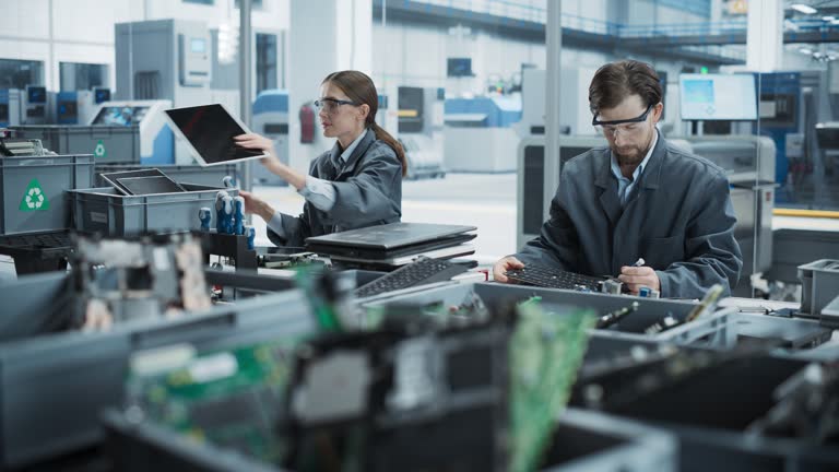 Caucasian Male And Female Workers Taking Apart Laptops To Recycle Electronic Components For Printed Circuit Board Production At Electronics Factory. Employees Unscrewing and Sorting Computer Parts