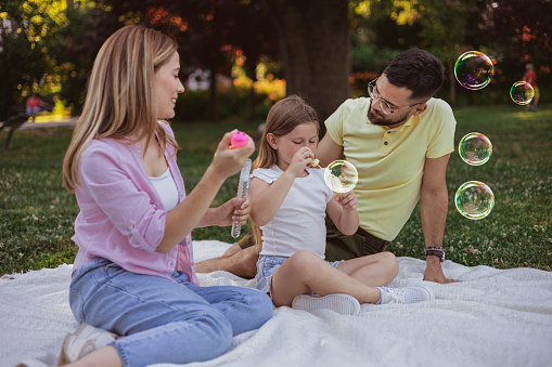 Happy Family With Child Girl Sitting on a Blanket while Blow Soap Bubbles Outdoor in Sunny Summer Day. Family, Leisure and People Concept