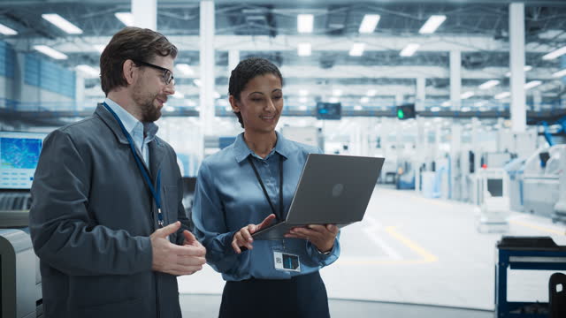 Caucasian Man and Hispanic Female Having a Conversation at an Industrial Facility. Advanced Factory Employees Working Together, Using Laptop Computer for Supervising Assembly Process Of New Devices.