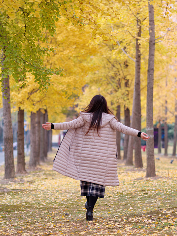 Rear view of Chinese girl in Long down jacket running with arms open enjoy carefree time in forest park in sunny day. Winter outdoor fashion portrait of glamour young Chinese woman.