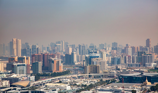 A sprawling cityscape of Dubai under a hazy sky. In the foreground, there are low-rise buildings with varied architectural designs, colors, and sizes. A major highway or expressway filled with vehicles stretches across the middle of the image, showcasing the city’s busy transportation network. Beyond the highway, there are more densely packed buildings including mid-rise and high-rise structures indicating a commercial or downtown area. In the distance, towering skyscrapers outline the city’s skyline.