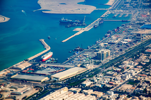 A bustling port area with docked ships, clear turquoise water, and surrounding urban landscape. The port is a hub of activity with industrial equipment and containers visible on the docks. The water is a clear turquoise color and the surrounding buildings include what appears to be residential and commercial structures. The roads adjacent to the port are visible with cars on them. The photograph showcases the vibrant urban landscape of Dubai UAE and the busy port area.