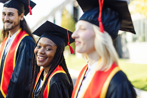 Man in a graduation gown holding a diploma and gesturing thumbs up in front of a USA flag