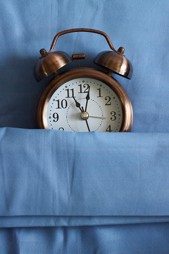 Stock photo showing close-up, elevated view of retro, double bell, analogue alarm clock tucked up for bedtime on pillow and under bedding. Healthy sleep routine concept.
