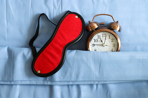 Stock photo showing close-up, elevated view of retro, double bell, analogue alarm clock tucked up for bedtime on pillow and under bedding with red eye mask. Healthy sleep routine concept.