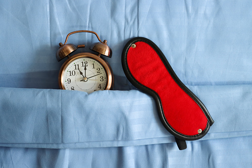 Stock photo showing close-up, elevated view of retro, double bell, analogue alarm clock tucked up for bedtime on pillow and under bedding with red eye mask. Healthy sleep routine concept.