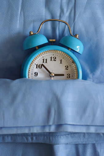 Stock photo showing close-up, elevated view of retro, double bell, analogue alarm clock tucked up for bedtime on pillow and under bedding. Healthy sleep routine concept.