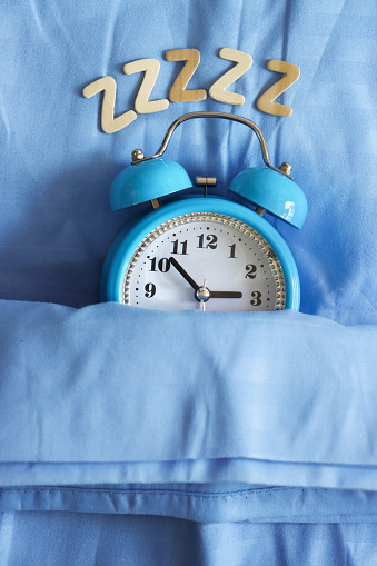 Stock photo showing close-up, elevated view of an arc of wooden letter z's above retro, double bell, analogue alarm clock with white dial tucked up for bedtime on pillow and under bedding. Healthy sleep routine concept.