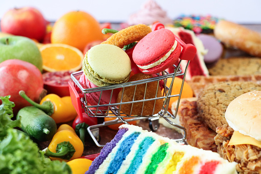 Stock photo showing close-up view of multi coloured macarons and chicken nuggets in a miniature, model shopping trolley surrounded by a of variety of sweet and savoury junk and healthy food items including red and green apples, chillies, pomegranate, watermelon quarters, red cabbage, macarons, rainbow and red velvet cake slices, chocolate bar, orange, chocolate chip cookies. Balanced diet concept.