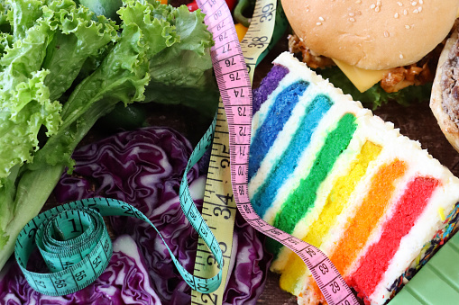 Stock photo showing close-up, elevated image of a variety of sweet and savoury junk and healthy food items including red cabbage, lettuce, chicken burger, rainbow cake slice, chocolate bar. Balanced diet concept.