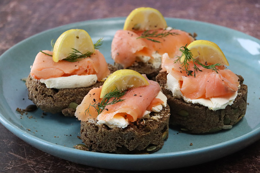 Stock photo showing close-up, elevated view of a teal blue plate containing an appetiser of four slices of dill bread topped with cream cheese and smoked salmon that have been garnished with fresh dill and a wedge of lemon.