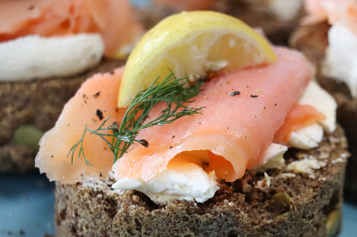 Stock photo showing close-up, elevated view of a teal blue plate containing an appetiser of four slices of dill bread topped with cream cheese and smoked salmon that have been garnished with fresh dill and a wedge of lemon.