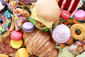 Full frame image of sweet and savoury junk foods, chicken burger, battered onion rings, pizza slices, glazed ring doughnuts, macarons, red velvet cake, croissant, cupcake with butter icing, fast food items, unhealthy eating concept, elevated view