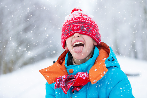 A funny little boy in blue winter clothes is walking during a snowfall. Winter outdoor activities for children. A cute child in a warm hat low above his eyes catches snowflakes with his tongue