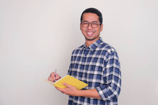 istock Adult Asian man smiling at the camera while writing something on a book 1887277010