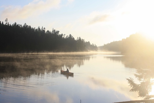 A solo canoeist paddles across calm waters as the sun rises over the misty horizon on a lake in the Canadian Wilderness.