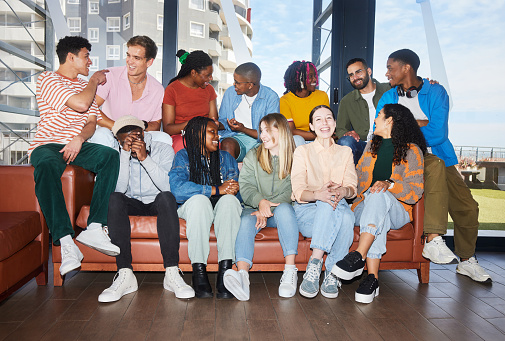 Diverse group of laughing young college students and their teacher sitting together on a sofa in a hallway at a school