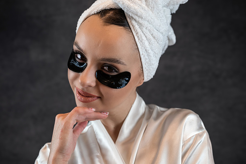 Attractive young woman applying collagen black eye patches under her eyes against a black wall.  Natural cosmetic for beauty care.