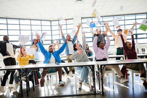 Diverse group of ecstatic young college students cheering and throwing the notes in the air in a classroom at the end of their school semester