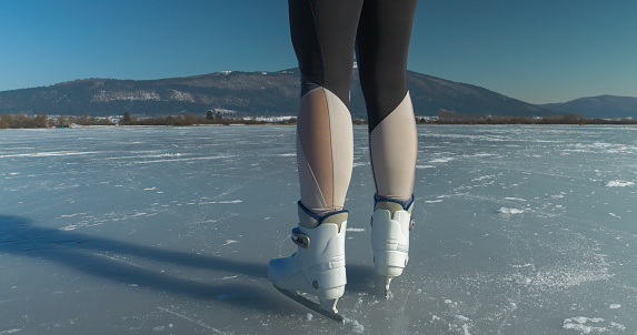 CLOSE UP: Female skater standing in the middle of a frozen lake in white ice skates. She is ice skating on a naturally frozen pond. A wonderful outdoor sports activity for cold and sunny winter days.
