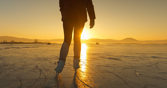 LENS FLARE, SILHOUETTE: A cold winter day with a woman skating on a frozen lake in golden sunset light. She skates across icy surface of a natural ice rink while last rays of sun peek over hills.