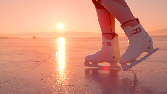 CLOSE UP, LENS FLARE: Female skates on glittering ice surface of a frozen lake at golden morning sunrise. Woman is gliding on naturally frozen ice with her white skates on a cold and sunny winter day.