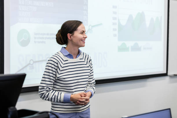 Young adult presenter listens to question during seminar Standing in front of the large projection screen, the young adult female speaker listens to a question during the seminar. projector screen classroom stock pictures, royalty-free photos & images
