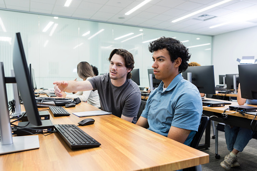 The young adult male tutor helps a young adult male university student with the assignment in the computer lab.
