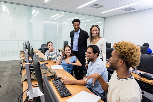 In the college computer lab, the mature adult male teacher and his fellow students listen to the young adult man.