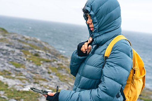 Latin woman trekking in an inhospitable area of the Irish coast wearing a hoodie and looking at a compass